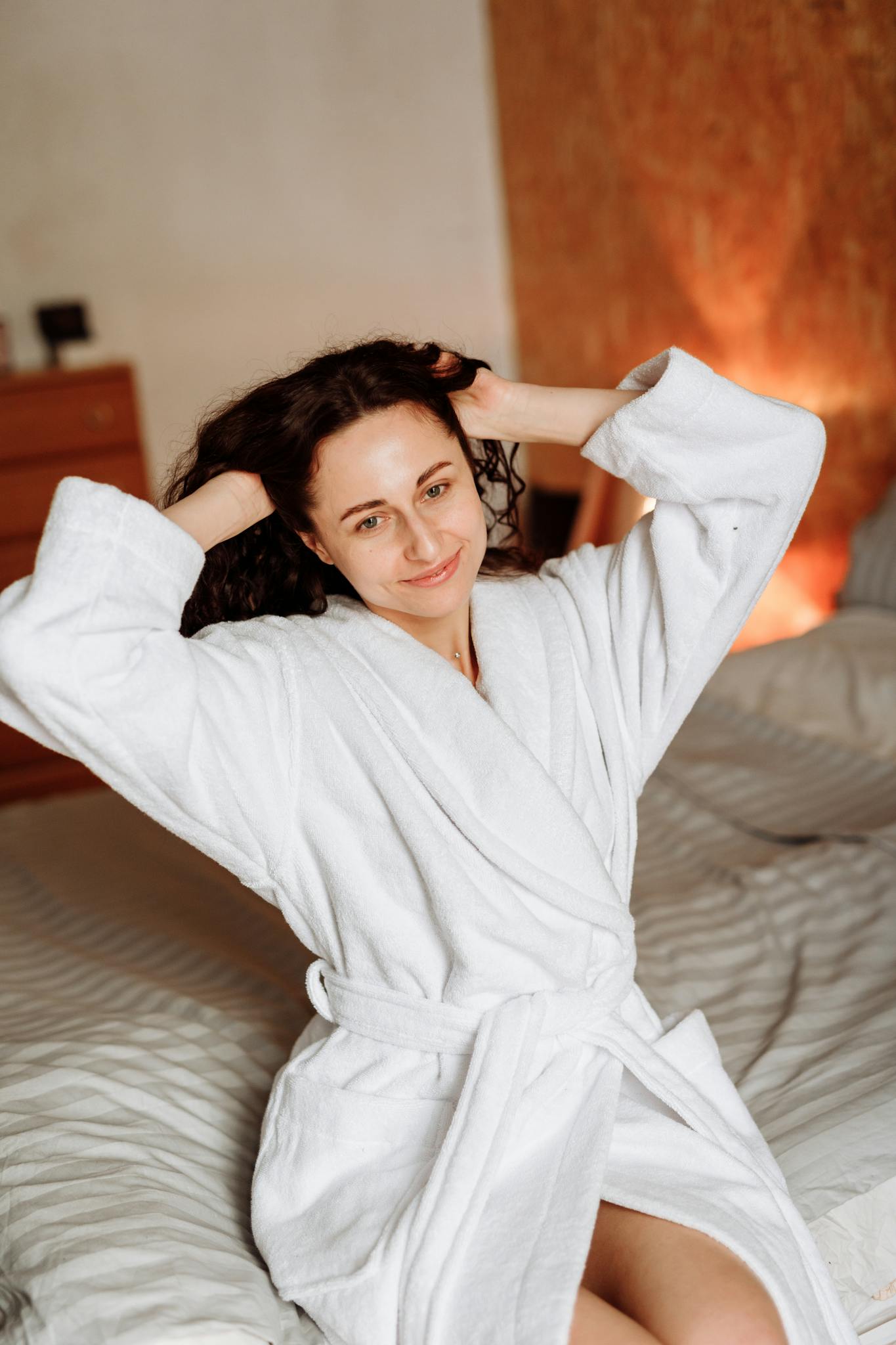 A young woman in a white bathrobe relaxes in a cozy bedroom setting.